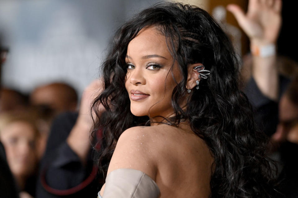 Rihanna, with long, wavy hair, wearing an ear cuff and an off-the-shoulder outfit, turns to face the camera at a celebrity event