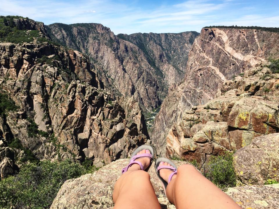 Two feet wearing purple sandals dangle off the edge of a steep cliff.