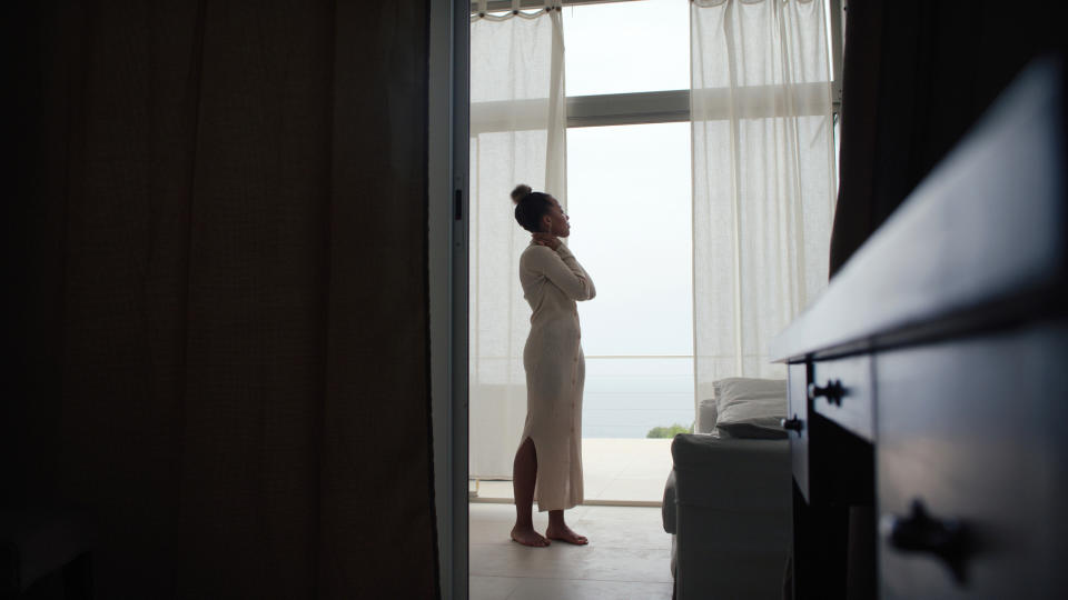Woman in a robe looking out a window, with a serene expression