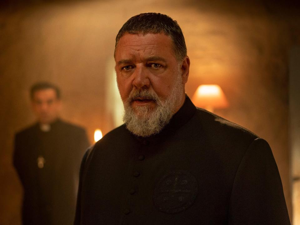 Russell Crowe as Father Gabriele Amorth stands in the foreground while another priest and a lamp are in the background.