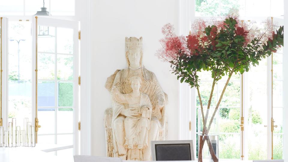 in a white dining room are two glass doors with an antique sculpture of the virgin and child against the wall between them, two geometric shaped white tables, three dining chairs with gray leather seats