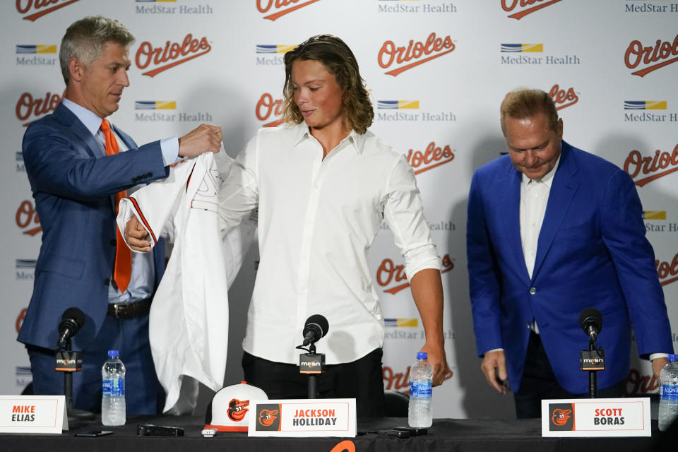 CORRECTS DATE - Jackson Holliday, center, the first overall draft pick by the Baltimore Orioles in the 2022 draft, gets a jersey from general manager Mike Elias, left, as agent Scot Boras looks on during a news conference introducing him to the Baltimore media prior to a baseball game between the Baltimore Orioles and the Tampa Bay Rays, Wednesday, July 27, 2022, in Baltimore. (AP Photo/Julio Cortez)