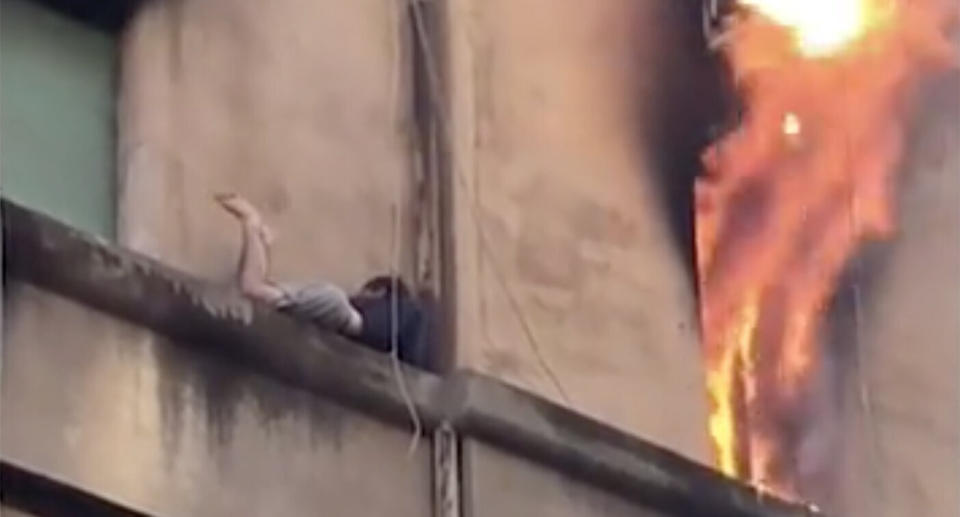 The young man clings to the narrow ledge while flames roar out of a nearby window.