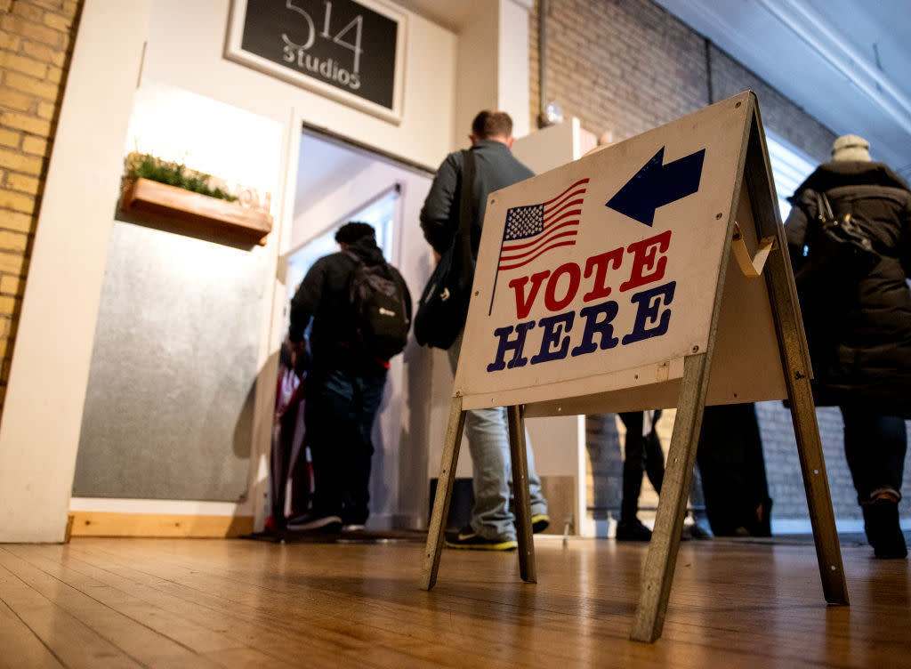 Voters lining up in a hall to vote. A sign with an American flag, an arrow and the text "Vote Here" is visible in the foreground.