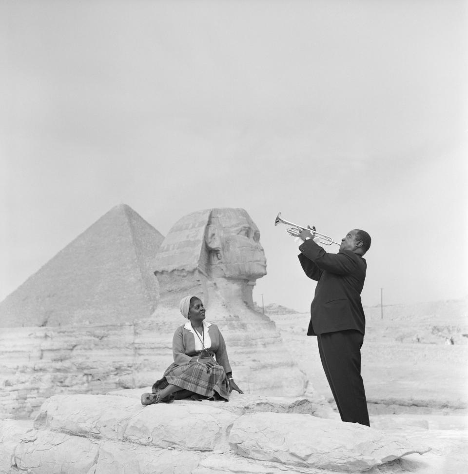 American jazz trumpeter Louis Armstrong plays the trumpet while his wife sits listening, with the Sphinx and one of the pyramids behind her, during a visit to the pyramids at Giza.