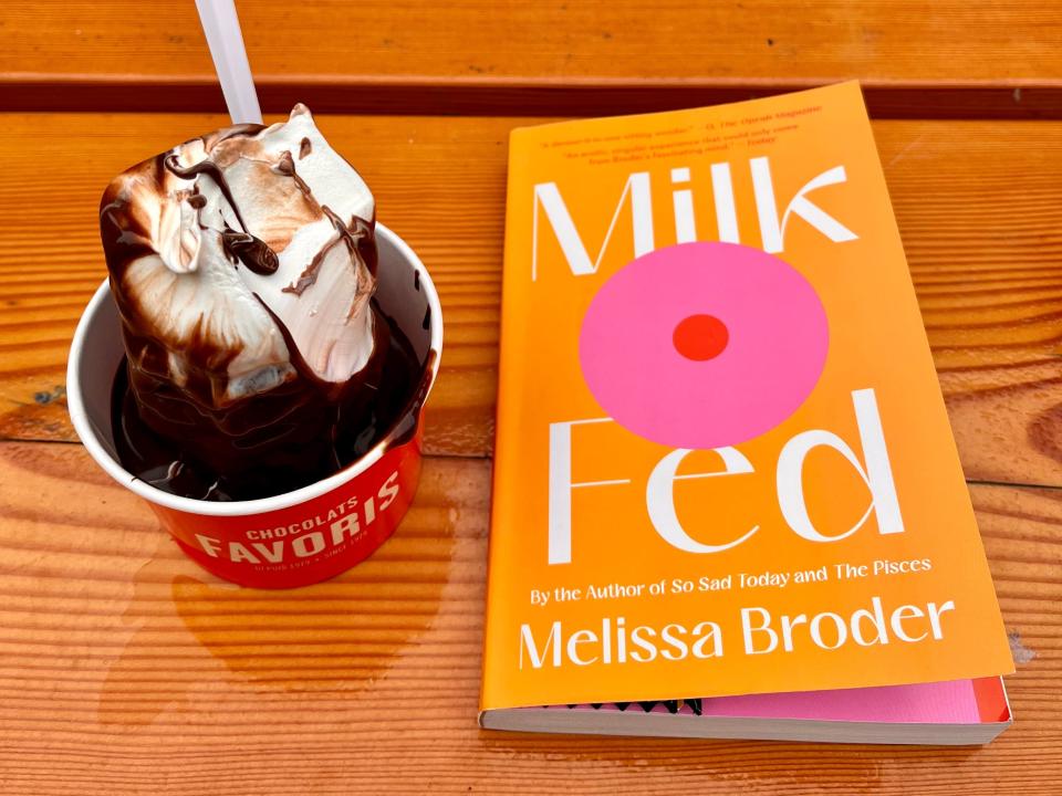 cup of ice cream with chocolate sauce next to a copy of milk fed novel on a table