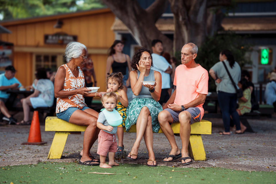 Family with two adults and two children eating on a bench in a public place with others around
