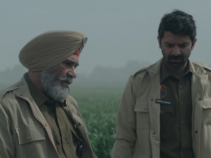 two men in kohrra, they are police officers wearing beige clothing and looking down at something in a field