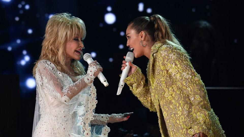 dolly parton and miley cyrus face each other while singing into microphones each of them holds, parton wears a white dress with a transparent beaded overlay, cyrus wears a green bedazzled coat