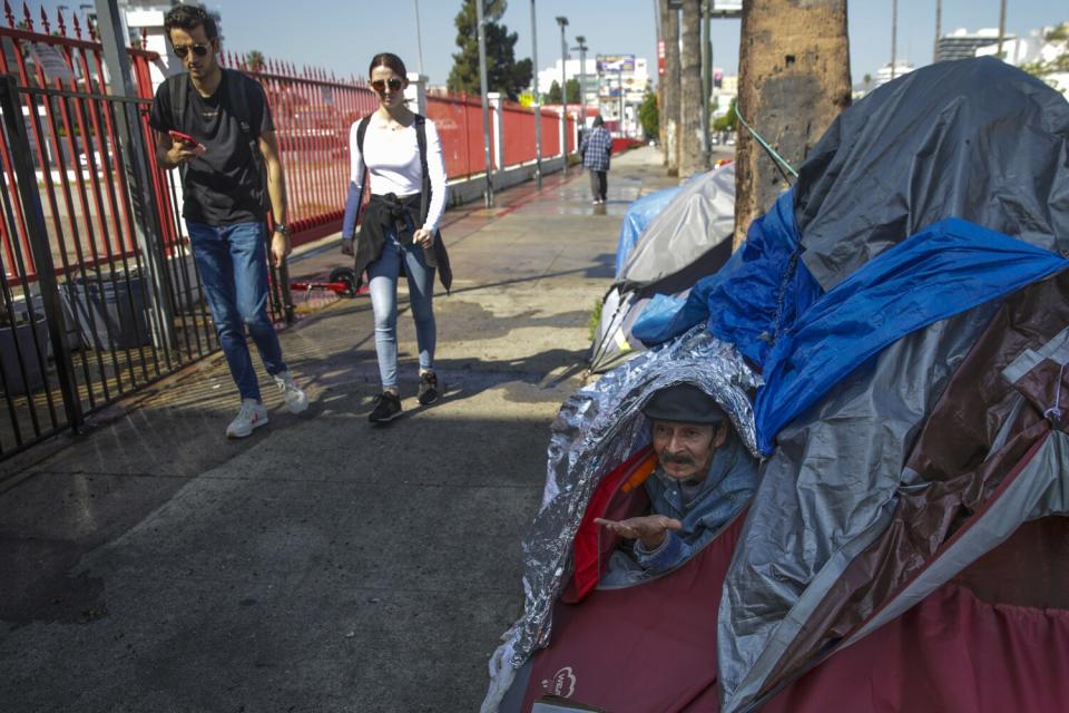 A man looks out from one of a row of tents as two pedestrians walk by.