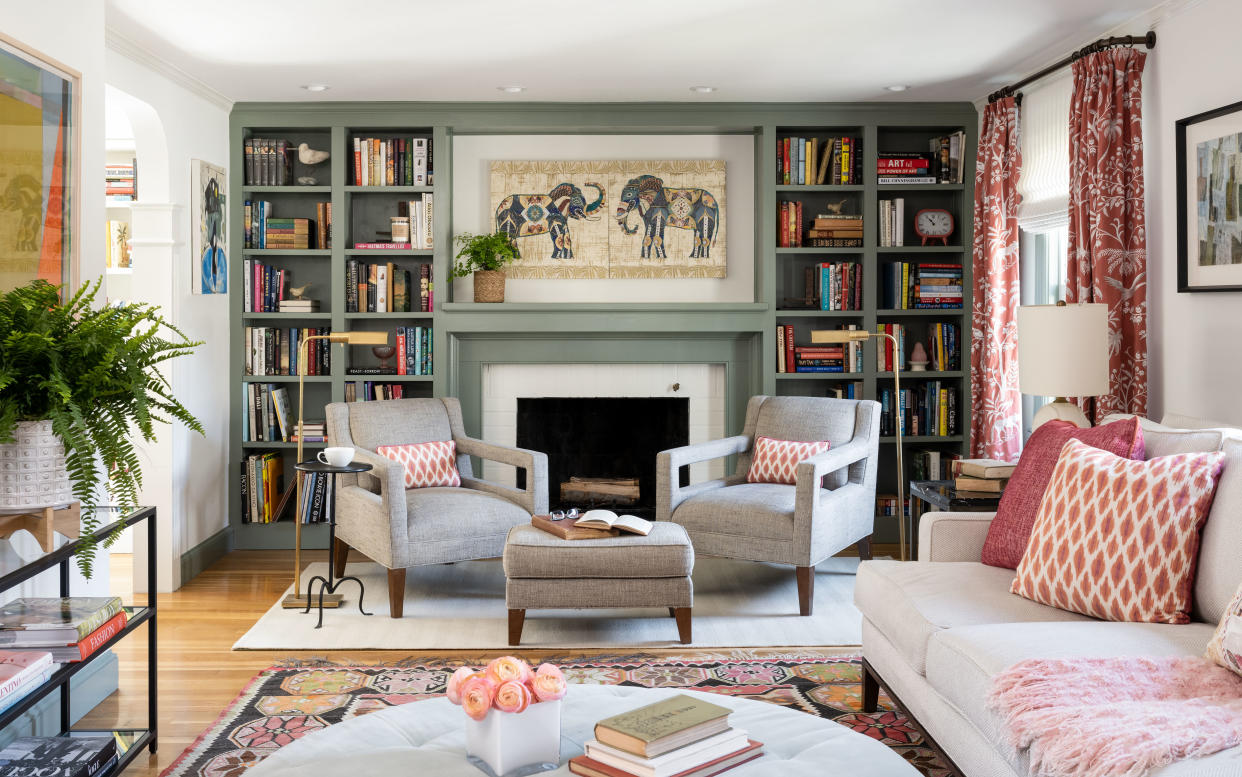  living room with green bookcase around fireplace, two armchairs, couch, rugs, floral drapes, pink cushions, footstool, plant 