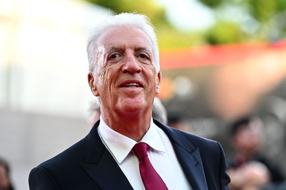 piero ferrari smiling for photos, he wears a navy blue suit jacket, white collared shirt and red tie