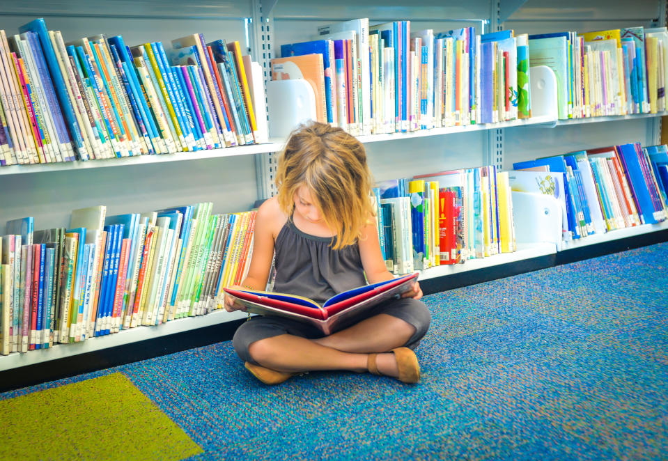 A young child sits on the floor reading a book in a library, surrounded by shelves filled with colorful books