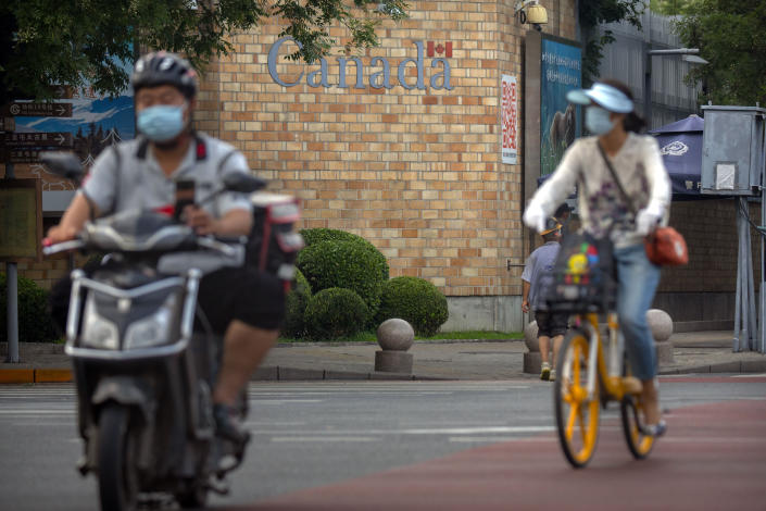 People wearing face masks to help protect against the coronavirus ride past the Canadian Embassy in Beijing, Thursday, Aug. 6, 2020. China has sentenced a Canadian citizen to death on charges of manufacturing the drug ketamine amid heightened tension between the two countries. (AP Photo/Mark Schiefelbein)