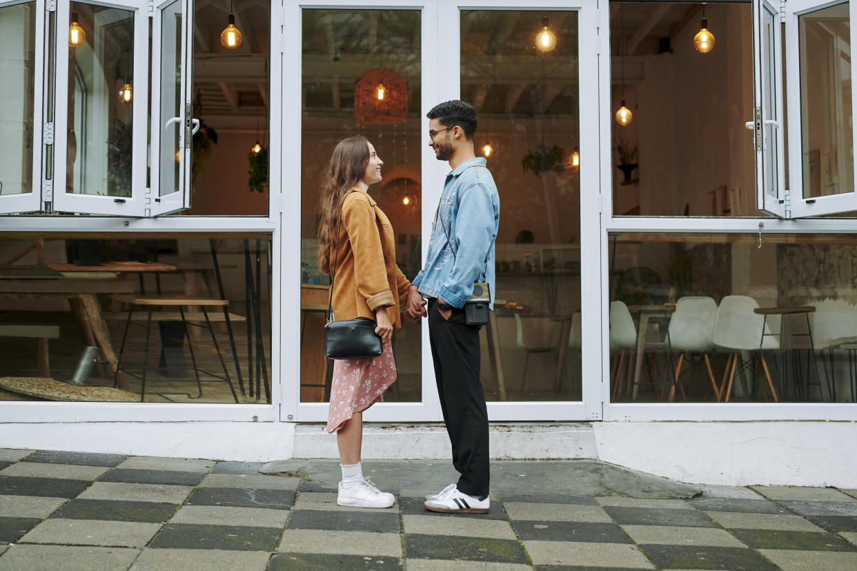 Smiling young couple holding hands together and standing face to face on a sidewalk outside of a cafe