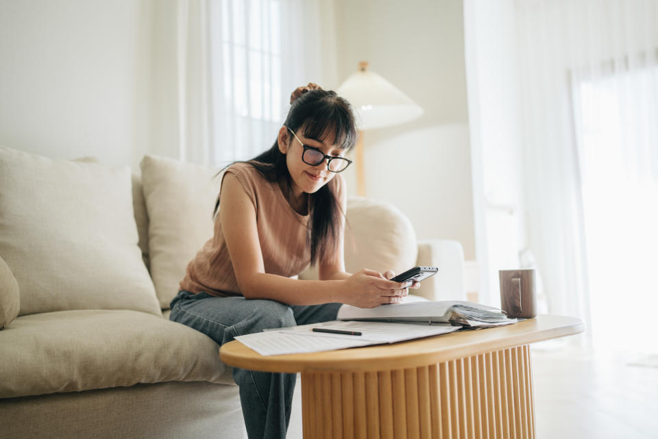 A woman sits at her living room with smartphone and financial reports doing her monthly budget. Looking for smart ways to save