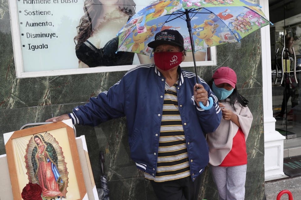 Un hombre y su hija, con mascarillas para protegerse del coronavirus, venden paraguas e imágenes de la Virgen de Guadalupe el miércoles 3 de junio de 2020, en Quito, Ecuador. (AP Foto/Dolores Ochoa)