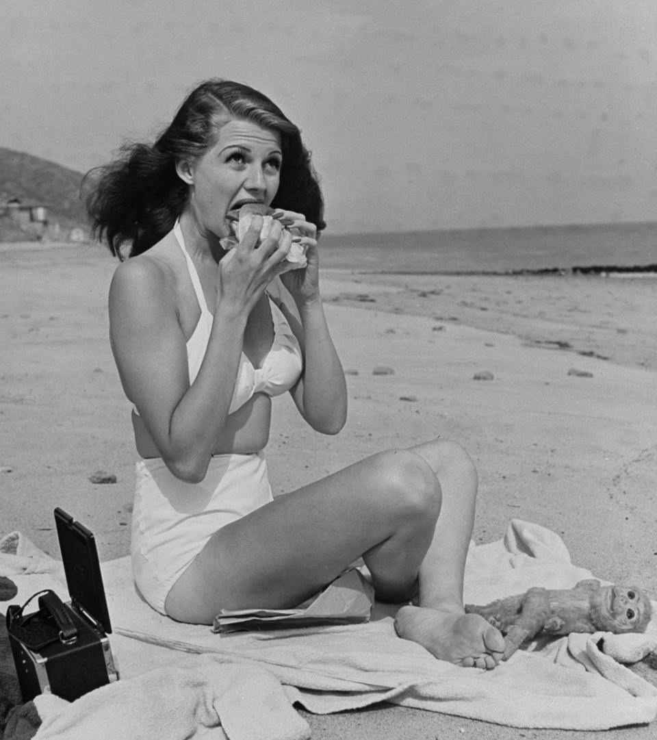 Taking a bite out of a hamburger as she sunbathes in a white two-piece bathing suit, circa 1941.