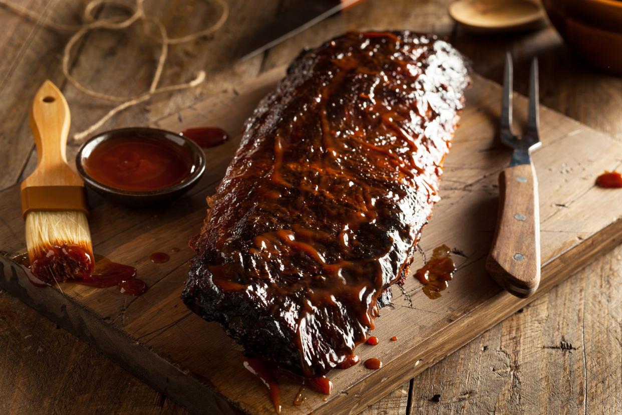 Bourbon-BBQ glazed baby back ribs on a wooden cutting board with utensils for BBQ and BBQ sauce surrounded by rustic items