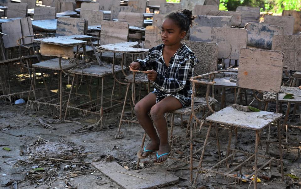 A girl sits in a muddy, destroyed school chair on muddy, messy ground