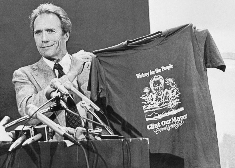 Clint holding up a "Clint Our Mayor" T-shirt at a podium