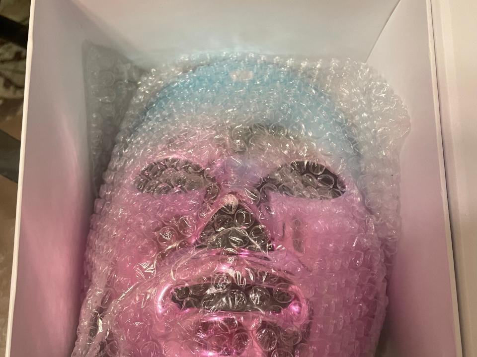 A purple and blue shiny face mask in a white box covered in bubble wrap.