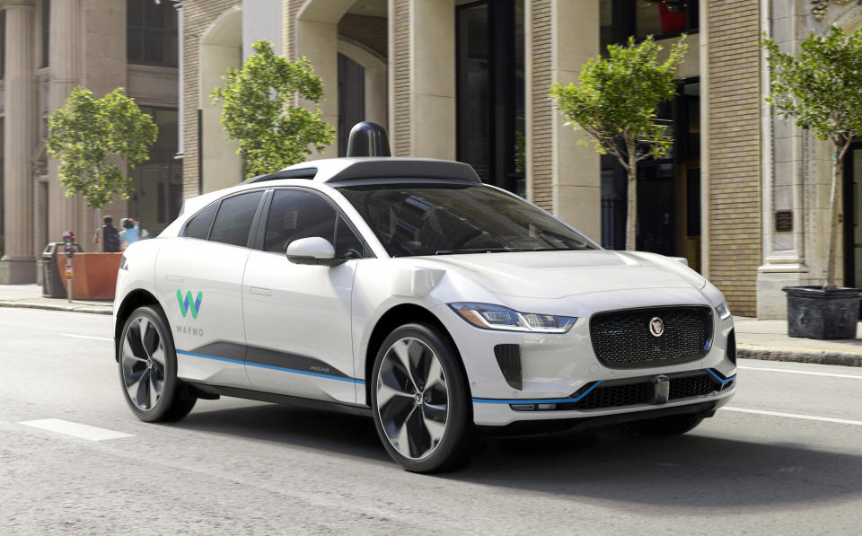 A white Jaguar I-Pace SUV, with self-driving sensor hardware and Waymo logos, on a city street.