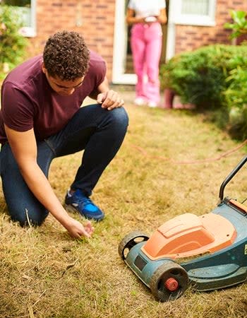 A man kneels to touch dry, brown grass next to a lawn mower. 