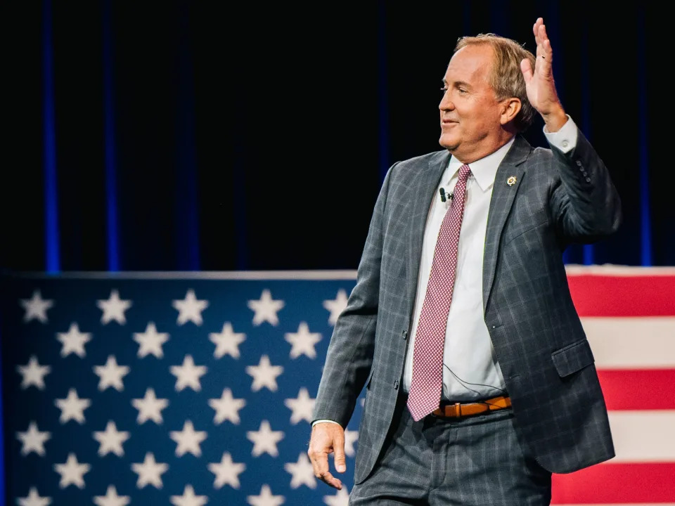 Texas Attorney General Ken Paxton waves in a suit in front of an oversized American flag