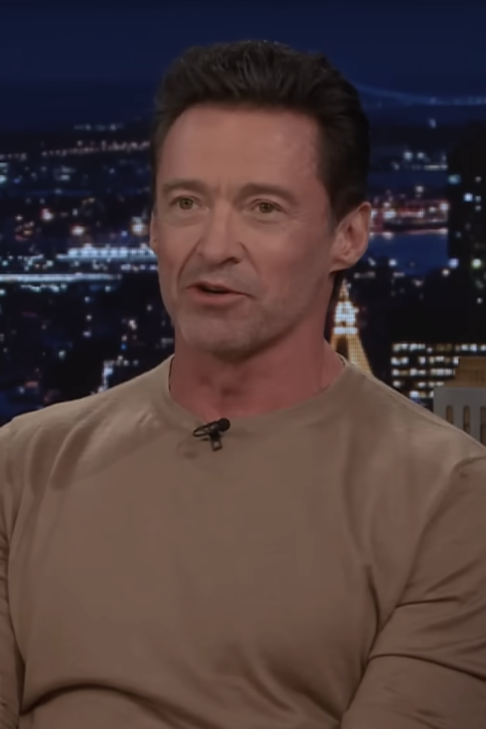 Hugh Jackman is speaking while seated against a background of a nighttime city skyline on a late-night talk show set