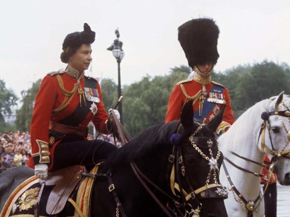 The Queen riding on a black horse in a red, black, and gold uniform with medals and a black bow-like headpiece.