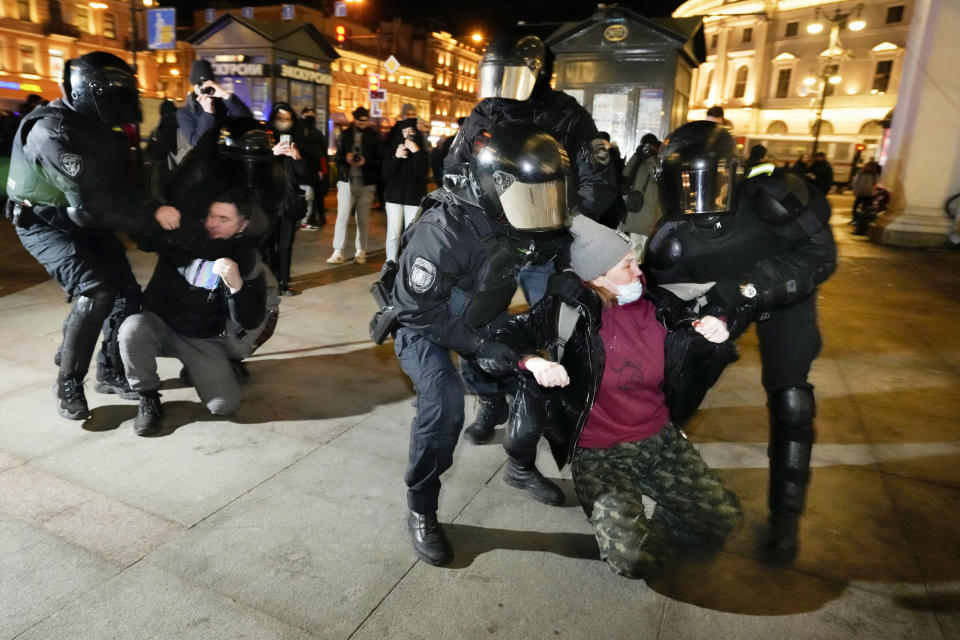 Police in face shields and riot gear forceably hold two kneeling people in a public square while others in the background train phones and cameras on them.