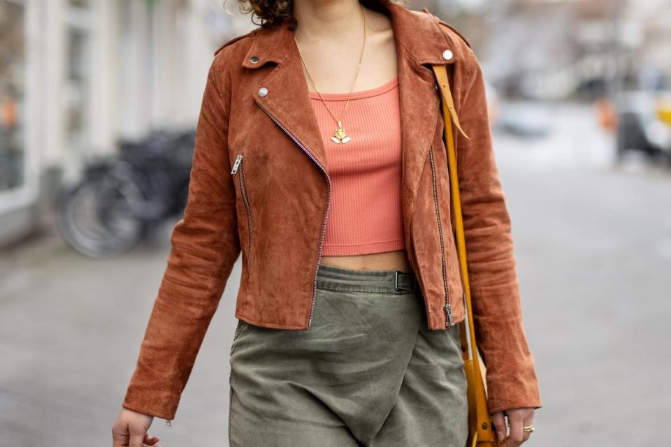 A woman with curly brown hair wearing a peach shirt, a tan motorcycle jacket, and olive bottoms walks down a city street.