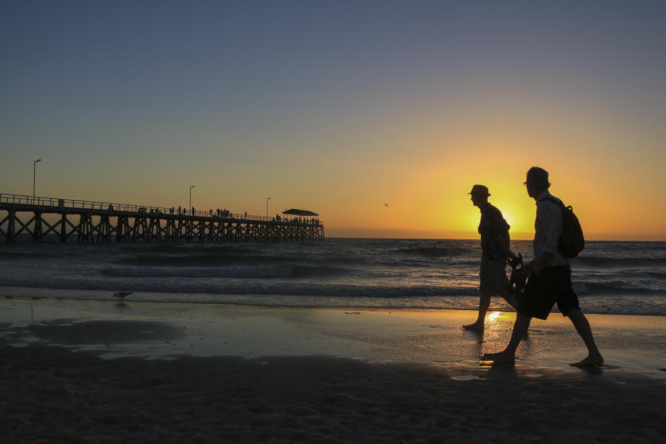 People walking on a beach with a jetty silhouetted against a setting sun in Adelaide, Australia.