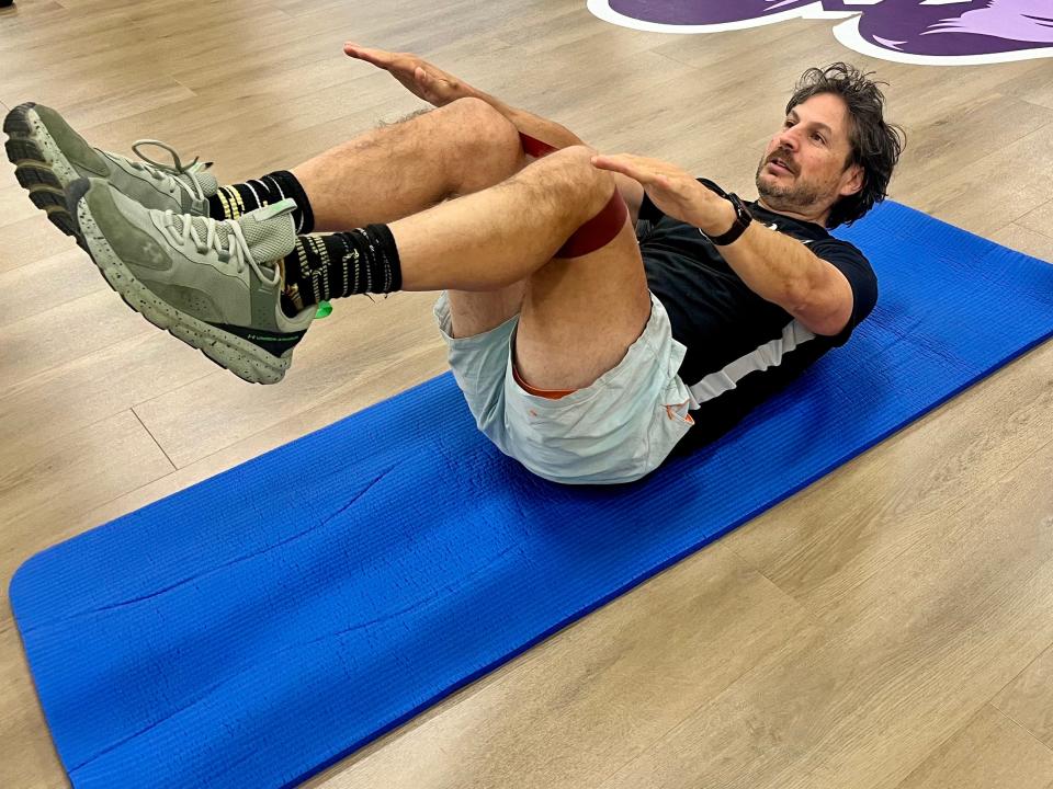 The writer lays on a blue Pilates mat with his legs bent at a 90-degree angle, toward his chest. He reaches his arms toward his knees