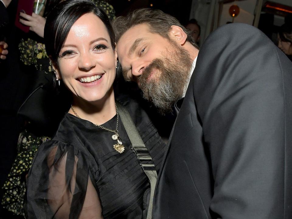 Lily Allen, in a black gown with sheer sleeves, smiles for photos while holding David Harbour, in a black suit.