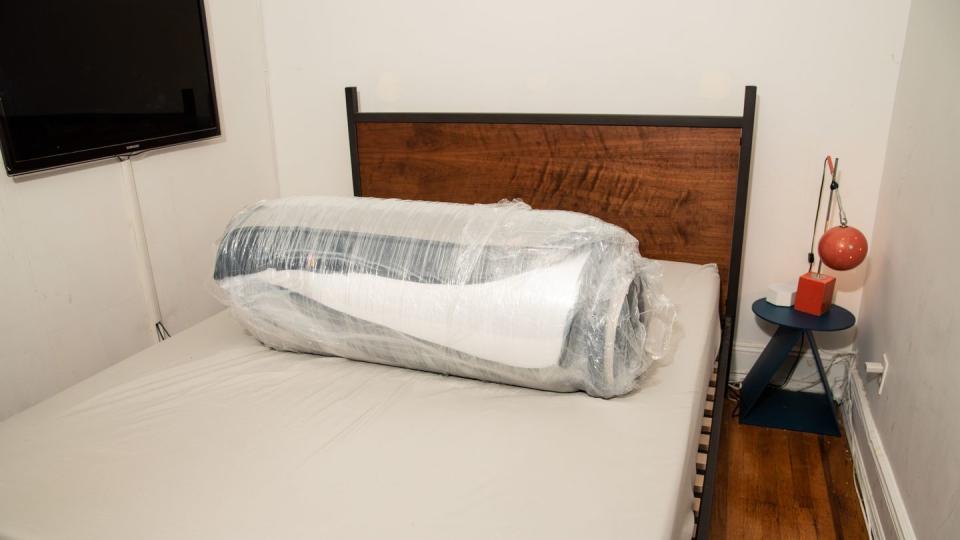 a compressed, rolled up mattress wrapped in plastic on a bed base