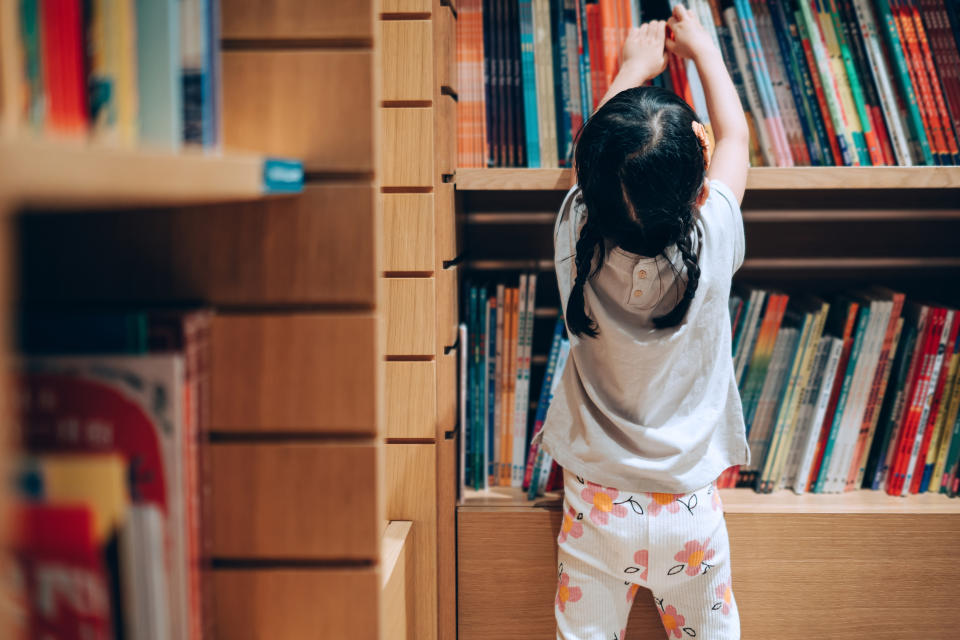 A child is reaching up to select a book from a shelf in a library. Shelves behind are filled with various books