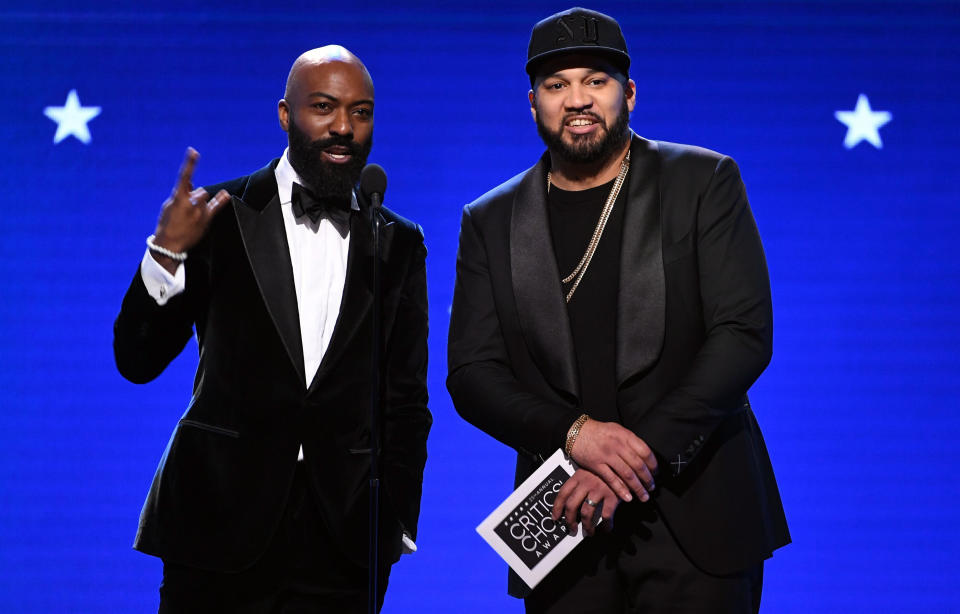Desus Nice and The Kid Mero in black suits while presenting at the critics choice awards in front of a blue backdrop