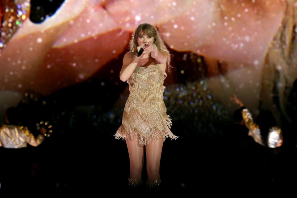 Taylor Swift pointing to the crowd as she performs on stage in a fringe dress