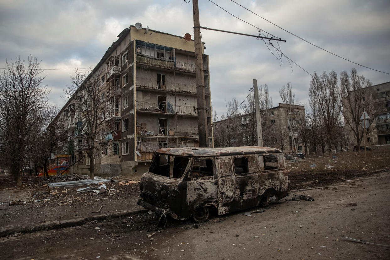 A burned-out van stands in front of a series of apartment blocks surrounded by litter.