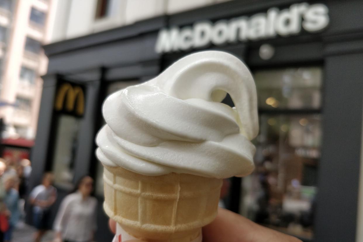 Person holding McDonald's soft serve ice cream cone with McDonald's restaurant in background.