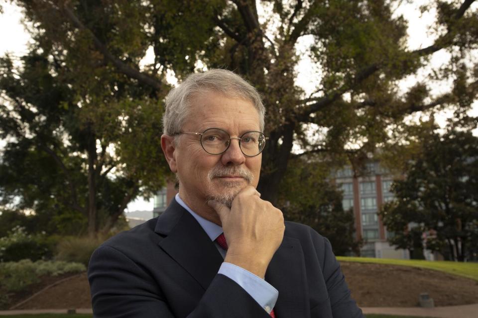 A man pictured from the shoulders up, his right hand on his chin, against a background of large trees.