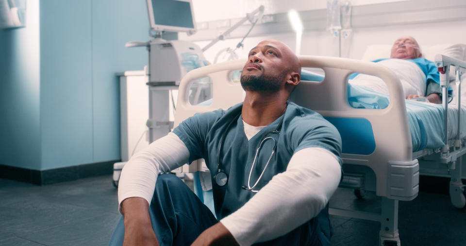 A healthcare worker in scrubs sits on the floor looking upward, appearing stressed. An elderly patient rests in a hospital bed in the background