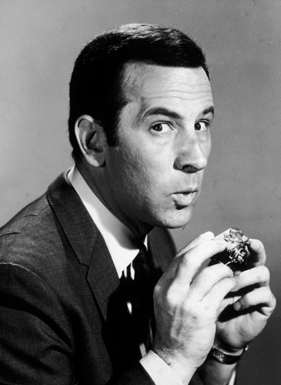 Portrait of American actor Don Adams, best known for his role as Maxwell Smart on the television series, 'Get Smart' (1965 - 1970), mugging while holding a miniature spy camera.