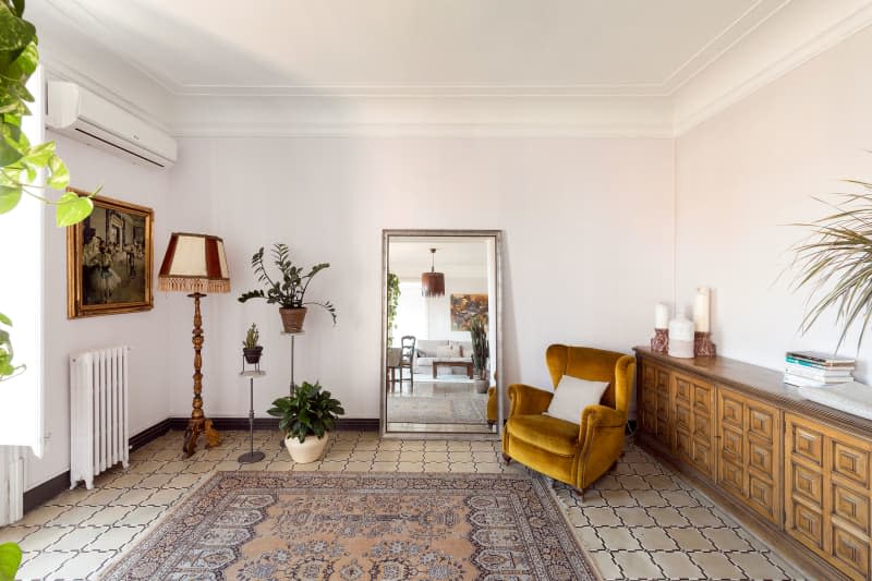 Wood storage unit lines wall of neutral toned living area decorated with mirror and plants.