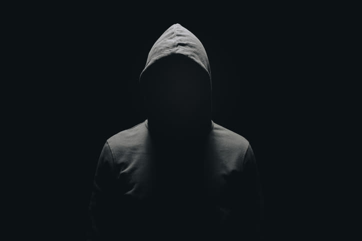 A person wearing a hooded sweatshirt stands in a dark, shadowy environment, with their face obscured by the hood