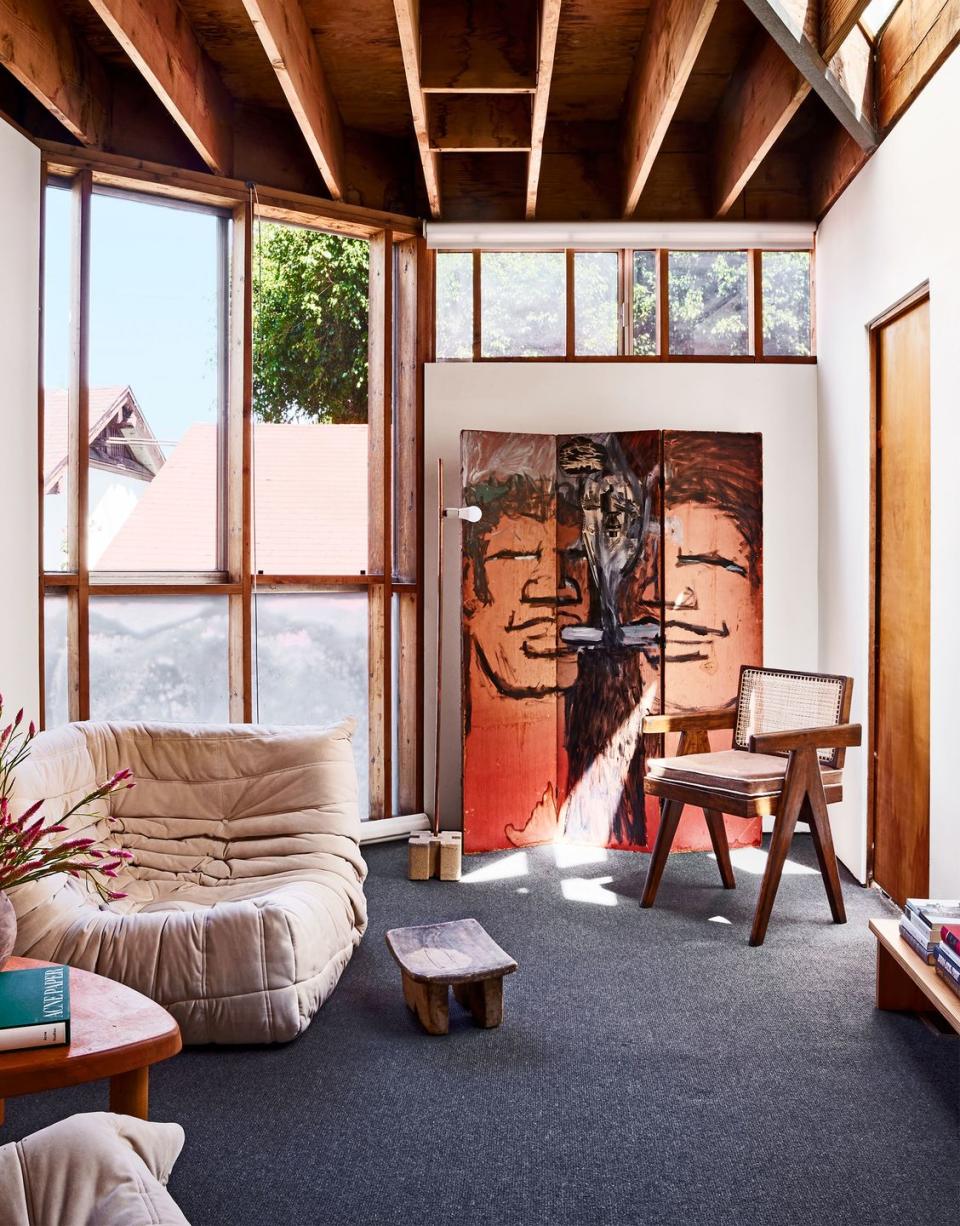 sitting room with two vintage armless lounge chairs in a taupe velvet, triangular wood cocktail table, vintage wood armchair with cane back and cushion seat, a folding screen with a cartoonish image of a man's face