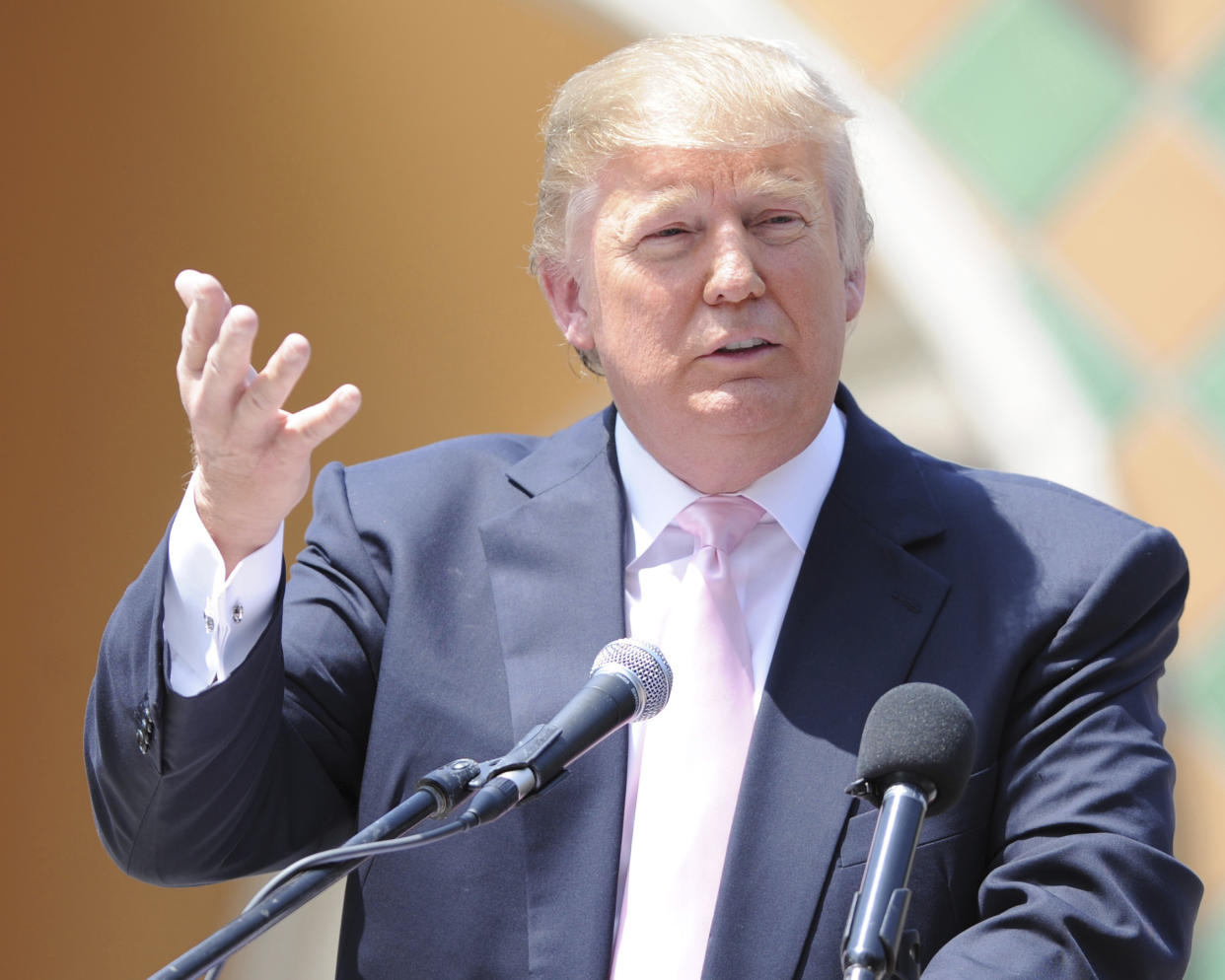  Donald Trump, wearing a pink tie with a dark blue suit, speaks with a raised right hand. 