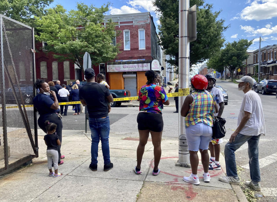 A cluster of people watch as Baltimore Police investigate a shooting, Wednesday, June 16, 2021, in Baltimore. One person was killed and five others were wounded Wednesday when gunmen walked up a street and opened fire on a Baltimore block from an intersection, the city's police commissioner said. (Kim Hairston/The Baltimore Sun via AP)